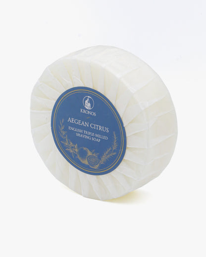 Kronos Aegean Citrus shaving soap turned at an angle on white, showing the depth of the puck and its pleated glassine wrap