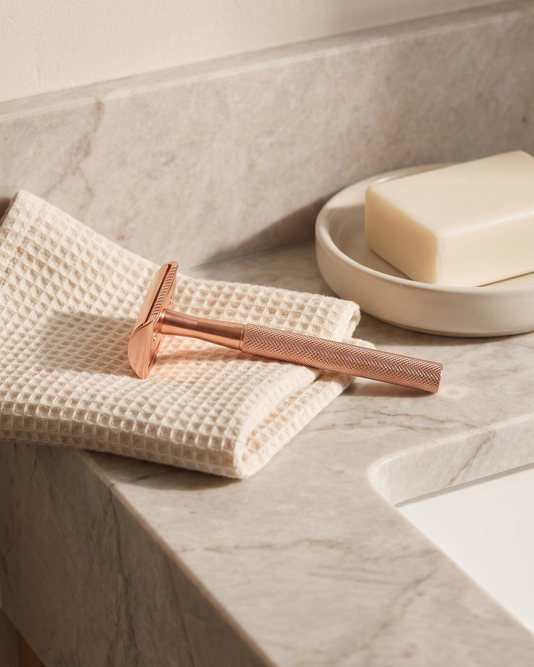 Rose gold razor resting on a waffle towel by a marble basin