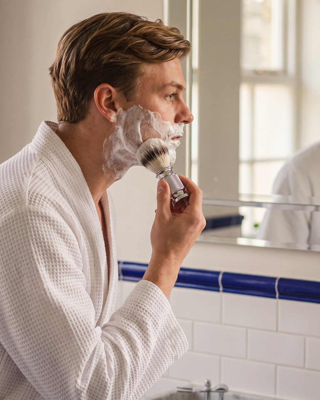 A man lathering his face with the Kronos shaving brush at a traditional bathroom basin