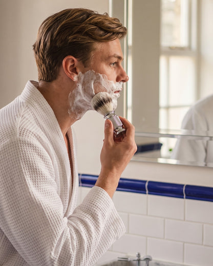 A man lathering his face with the Kronos shaving brush at a traditional bathroom basin